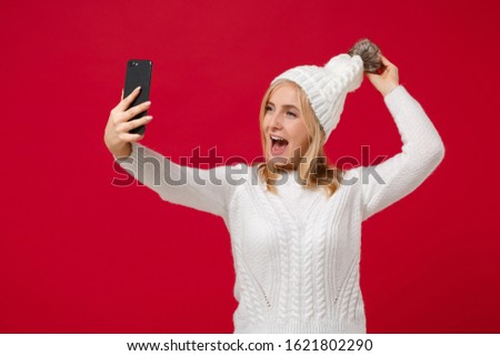 Cheerful funny young woman in white sweater, hat isolated on red wall background studio portrait. Healthy fashion lifestyle, cold season concept. Mock up copy space. Doing selfie shot on mobile phone