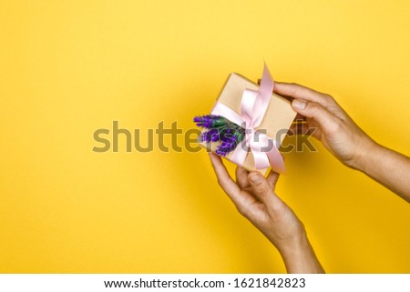 close-up woman hands holding a gift box with a lavender twig in a gesture of giving on yellow background with space