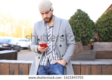 Handsome young man in grey coat and hat using smartphone, resting, standing on city street. Urban concept.