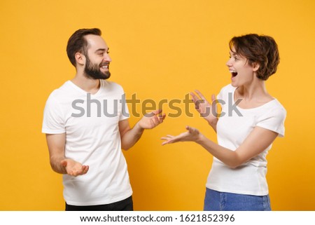 Funny young couple friends bearded guy girl in white t-shirts posing isolated on yellow orange background. People lifestyle concept. Mock up copy space. Looking at each other speaking spreading hands