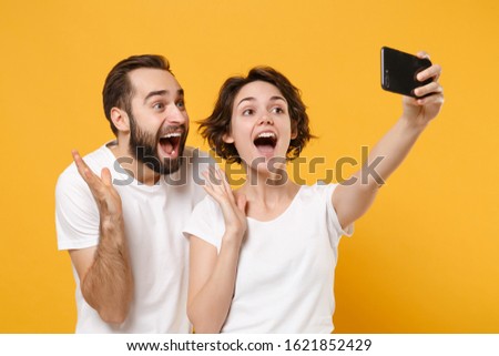 Excited young couple friends bearded guy girl in white t-shirts isolated on yellow orange background. People lifestyle concept. Mock up copy space. Doing selfie shot on mobile phone, spreading hands