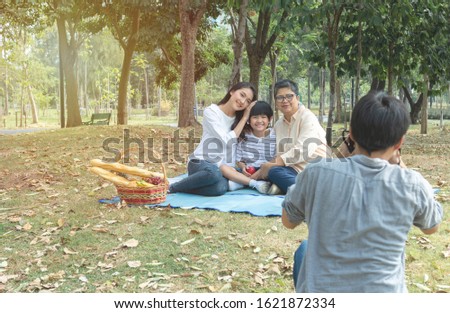 Asian father use digital camera take photo of his wife and son and grandma in park.Leisure time of asia family have picnic in park and take group photo together.