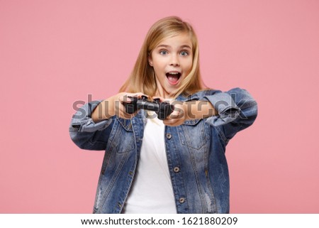 Excited little blonde kid girl 12-13 years old in denim jacket posing isolated on pastel pink background children portrait. Childhood lifestyle concept. Mock up copy space. Play game with joystick