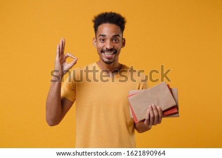 Smiling young african american guy in casual t-shirt posing isolated on yellow orange wall background. People lifestyle concept. Mock up copy space. Hold in hands books, notebooks showing OK gesture