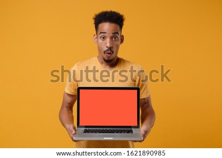 Shocked young african american guy in casual t-shirt posing isolated on yellow orange wall background. People lifestyle concept. Mock up copy space. Hold laptop pc computer with blank empty screen