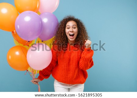Happy african american girl in orange knitted clothes isolated on pastel blue background. Birthday holiday party, people emotions concept. Celebrating hold colorful air balloons doing winner gesture