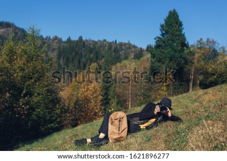 girl photographer with backpack lies on slope and shoot wildlife in mountains in autumn.