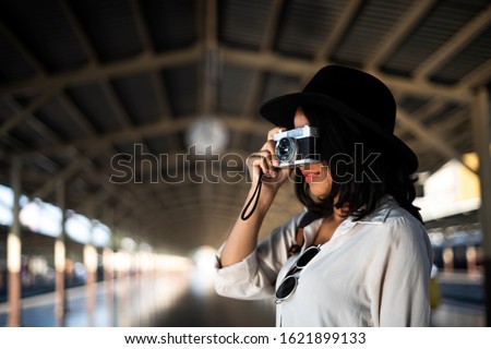 Wavy hair woman in white shirt and black hat take a photo with one hand while standing in the hallway of train platform.