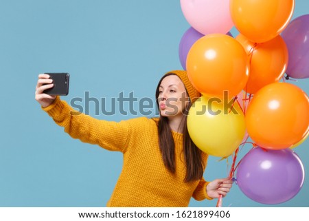 Pretty woman in sweater hat posing isolated on blue background. Birthday holiday party, people emotions concept. Mock up copy space. Celebrating hold air balloons doing selfie shot on mobile phone