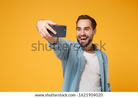 Funny young man in casual blue shirt posing isolated on yellow orange wall background studio portrait. People sincere emotions lifestyle concept. Mock up copy space. Doing selfie shot on mobile phone
