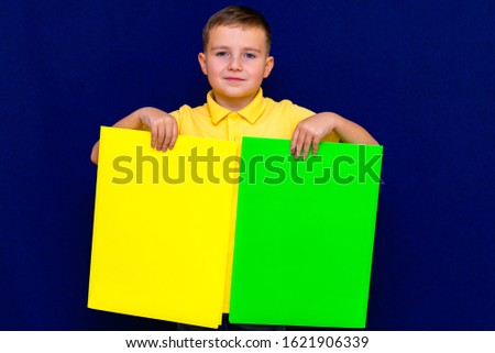 Cunning caucasian blue-eyed  boy  in yellow t-shirt holds light yellow and green blank cardboards on blue studio background. joyful schoolboy holding advertising sheet, copy space.