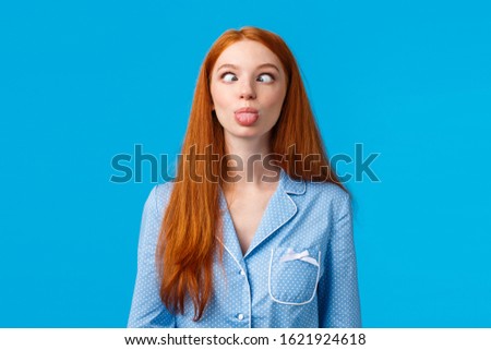 Be funny and silly. Redhead carefree and enthusiastic caucasian girl squinting, making goofy expression, showing tongue, standing blue background in nightwear, fool around