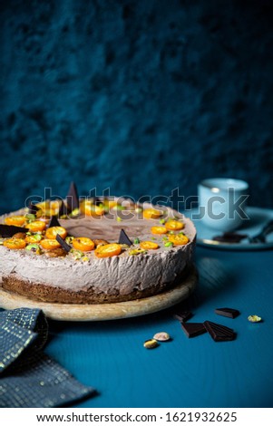 The whole layered chocolate mousse cake with orange jam, kumquats and pistachios on wooden board, cup of coffee and kitchen towel  on peacock blue wooden surface with dark background.