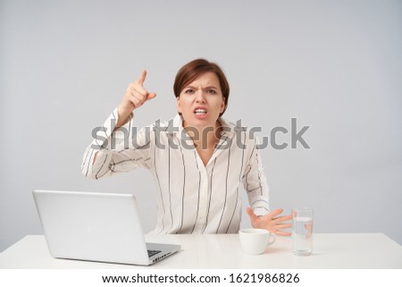 Angry young brown-eyed short haired woman with natural makeup raising excitedly forefinger while looking heatedly at camera, sitting at table over white background