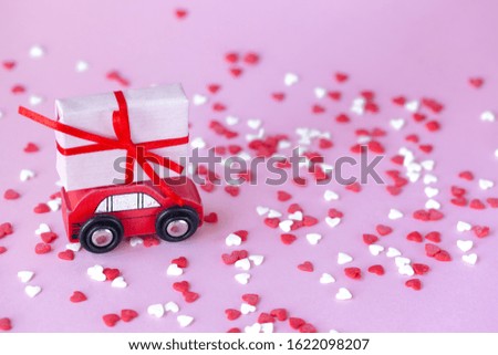 Valentines day delivery concept. Miniature red car with a present box on the roof on a pink background with red and wihte hearts