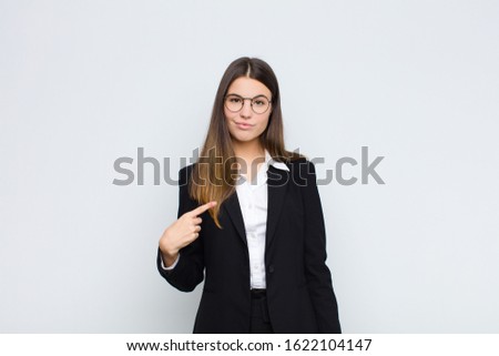 young businesswoman looking proud, confident and happy, smiling and pointing to self or making number one sign against white wall