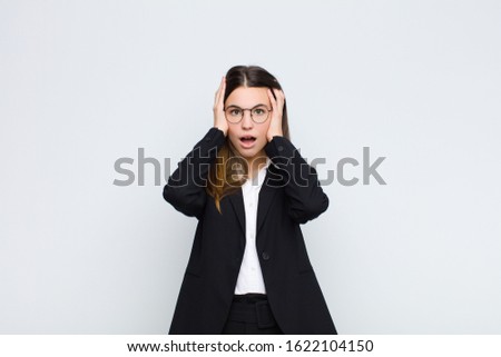 young businesswoman looking unpleasantly shocked, scared or worried, mouth wide open and covering both ears with hands against white wall