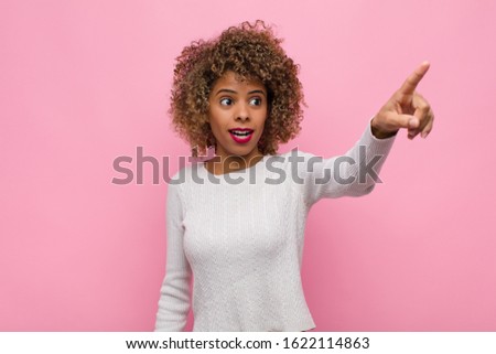 young african american woman feeling shocked and surprised, pointing and looking upwards in awe with amazed, open-mouthed look against pink wall