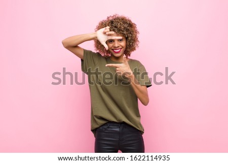 young african american woman feeling happy, friendly and positive, smiling and making a portrait or photo frame with hands against pink wall