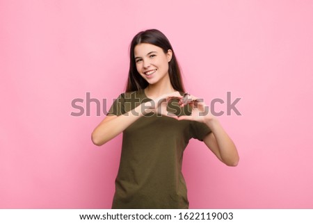 young pretty woman smiling and feeling happy, cute, romantic and in love, making heart shape with both hands against pink wall
