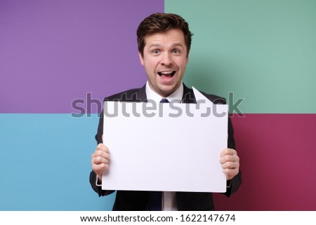 Young caucasian businessman in business outfit holding at blank canvas over colored background