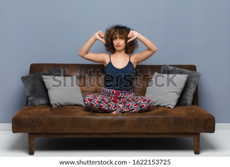 young pretty woman looking sad, disappointed or angry, showing thumbs down in disagreement, feeling frustrated sitting on a sofa.