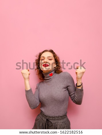 Portrait of joyful curly girl rejoices in victory with raised hands on pink background, looks in cameras and smiles. Happy joyful lady with red hair isolated on pink background. Copy space. Vertical