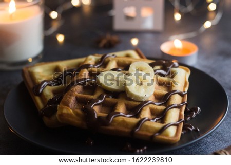 Viennese waffles on a plate with banana and chocolate. In the background are lights and candles. Romantic picture