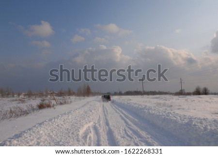 Winter sunny landscape on which an off-road car rushes along a snow-covered road to the horizon and white-blue clouds, leaving behind snow dust and long blue furrows from tires.