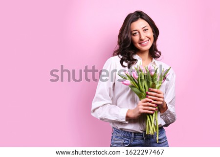 Studio portrait of gorgeous young brunette woman with long wavy hair wearing white loose cotton shirt, holding bouquet of tulip flowers. Pink isolated background, copy space, close up.