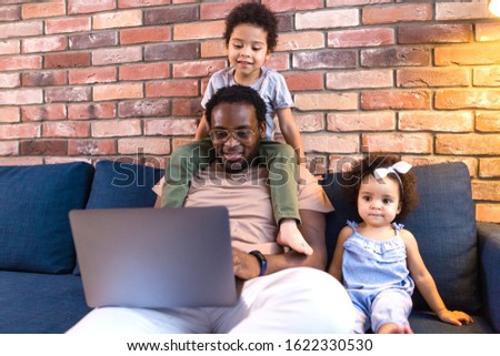 African dad with two children sitting on the sofa in the living room watching cartoons on a laptop. Son sitting on the neck of the Pope
