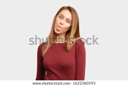 Funny girl makes faces sticking her tongue. The concept of frivolous fun and teasing. Beautiful young woman with long hair, clean smooth skin wears Burgundy-colored jacket sleeves isolated background