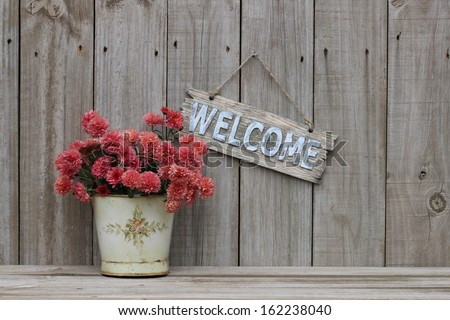 Wood welcome sign with pot of pink spring flowers - mums - hanging on distressed old rustic wooden fence; Mothers Day and floral background with copy space