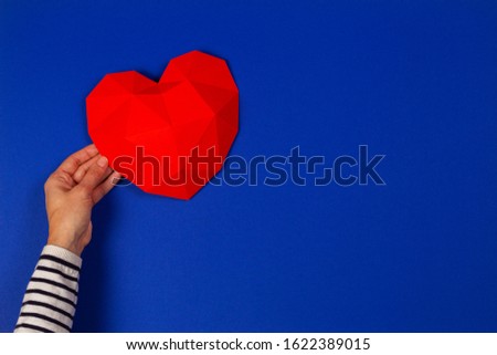 Female hand holding red polygonal heart on blue background. Top view