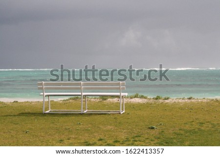 image of a white bench shown just to left of centre of the picture. bench is placed on green grass looking out over a blue ocean with a stormy grey sky.