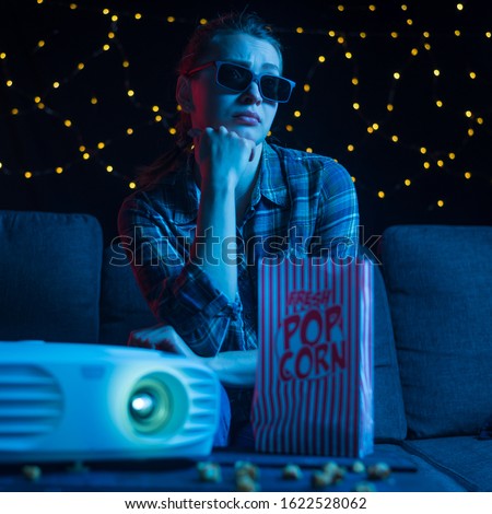 A young girl in 3D glasses watches a movie or a movie with a projector on the sofa with popcorn. Home relaxation, home cinema. Against the background with lights. Square photo