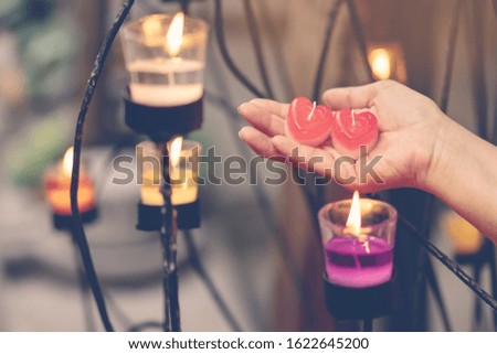 A red heart shaped candle in a woman's hand with candlelight in many small glass cups. A valentine's day background.