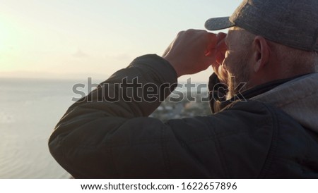 Senior traveler making binocular gesture and looking landscape sitting on a hill