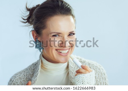 Portrait of smiling elegant woman in roll neck sweater and cardigan with lip balm as winter lip care isolated on winter light blue background.