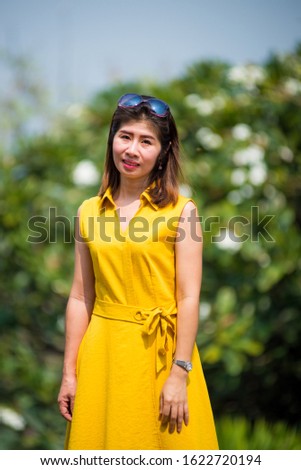 A woman is wearing a yellow dress. There is a sunblock on the head. Standing in front of the outdoor Half body shot, background is a green tree.