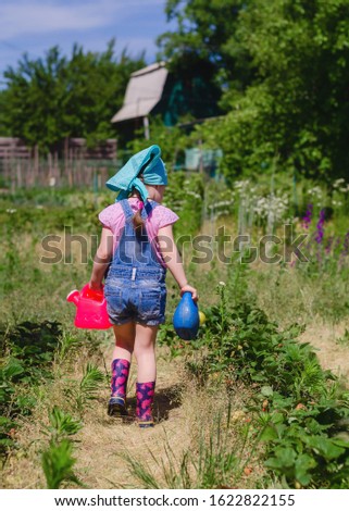 girl 4 years old, dressed in denim overalls, rubber boots and a bandana on his head watered strawberries from watering cans in the garden. Gardening. Spring, the beginning of summer.