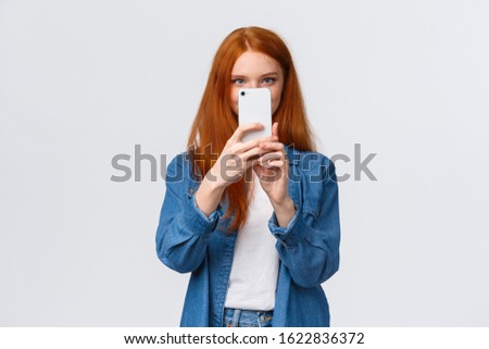 Turning on photo filter. Cheerful and cute female redhead lifestyle blogger, taking picture on smartphone, holding mobile phone near face, taking good shot, searching pretty angle, white background