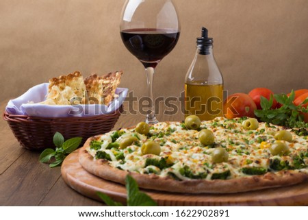 Broccoli pizza served on wooden board. Baked with mozzarella, cream cheese, broccoli, corn, peas, olives and sauce. Crostini, olive oil, basil, glass of red wine and tomatoes. Horizontal photography.