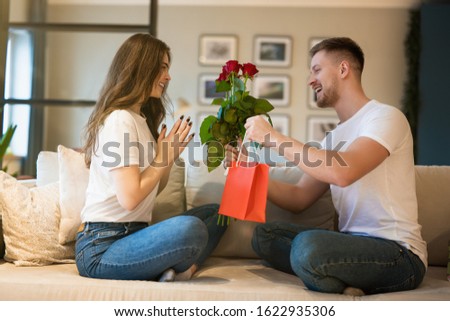 young lovely couple sitting on the sofa, man surprising wife with flowers and gift on saint valentine's day, happy unniversary