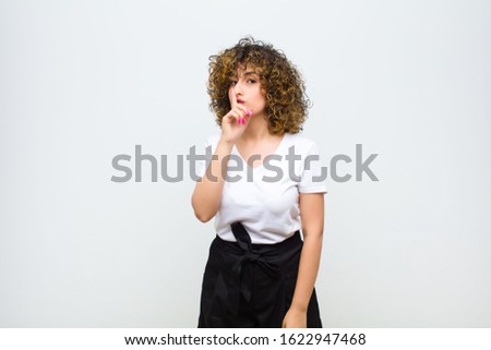 young pretty woman asking for silence and quiet, gesturing with finger in front of mouth, saying shh or keeping a secret against white wall