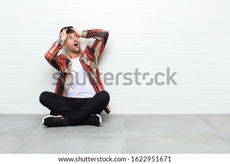 young handsome man with open mouth, looking horrified and shocked because of a terrible mistake, raising hands to head sitting on cement floor