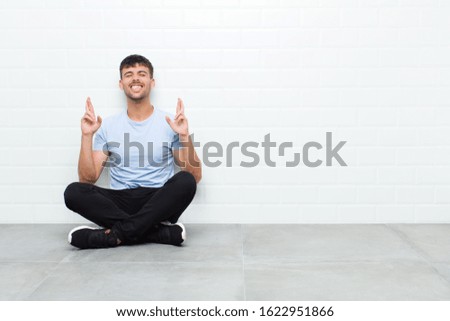 young handsome man smiling and anxiously crossing both fingers, feeling worried and wishing or hoping for good luck sitting on cement floor