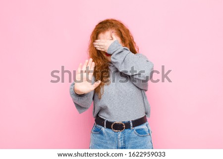 young red head woman covering face with hand and putting other hand up front to stop camera, refusing photos or pictures against pink wall