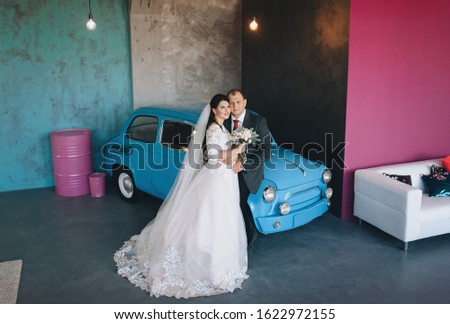 Stylish groom and beautiful brunette bride in a white dress with a bouquet are hugging in a modern studio on a background of a retro car. Wedding portrait of happy newlyweds. Photography, concept.