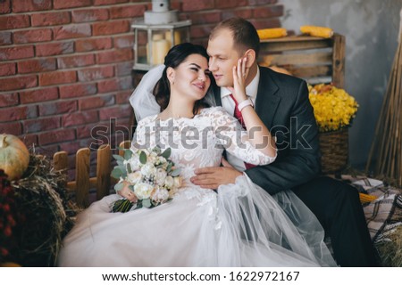 Stylish groom and beautiful bride in a white dress with a bouquet are sitting on the hay in the studio with autumn decor. Wedding portrait of happy and smiling newlyweds. Photography, concept.